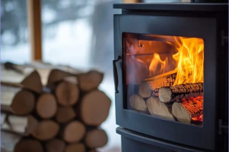 Log burner installation by Swept. Huddersfield A wood-burning stove with flames and stacked firewood in a winter setting.